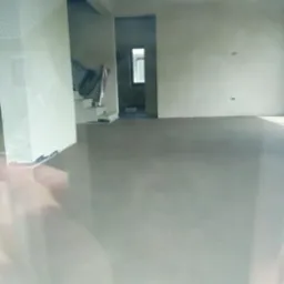 Wnętrze w stanie surowym z wylaną, szarą posadzką, widoczne ściany przygotowane do malowania i okno w oddali, częściowo rozmazane odbicie w szybie.