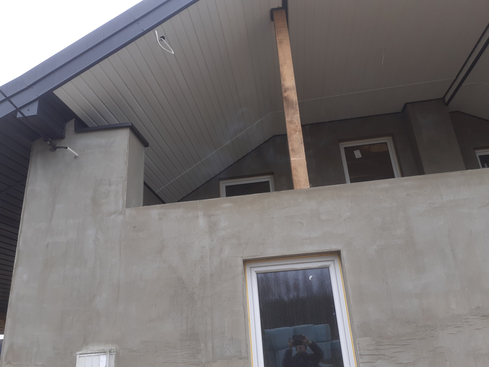 Surowa ściana budynku z widoczną drewnianą podporą dachu, okna z odbiciem osoby fotografującej.