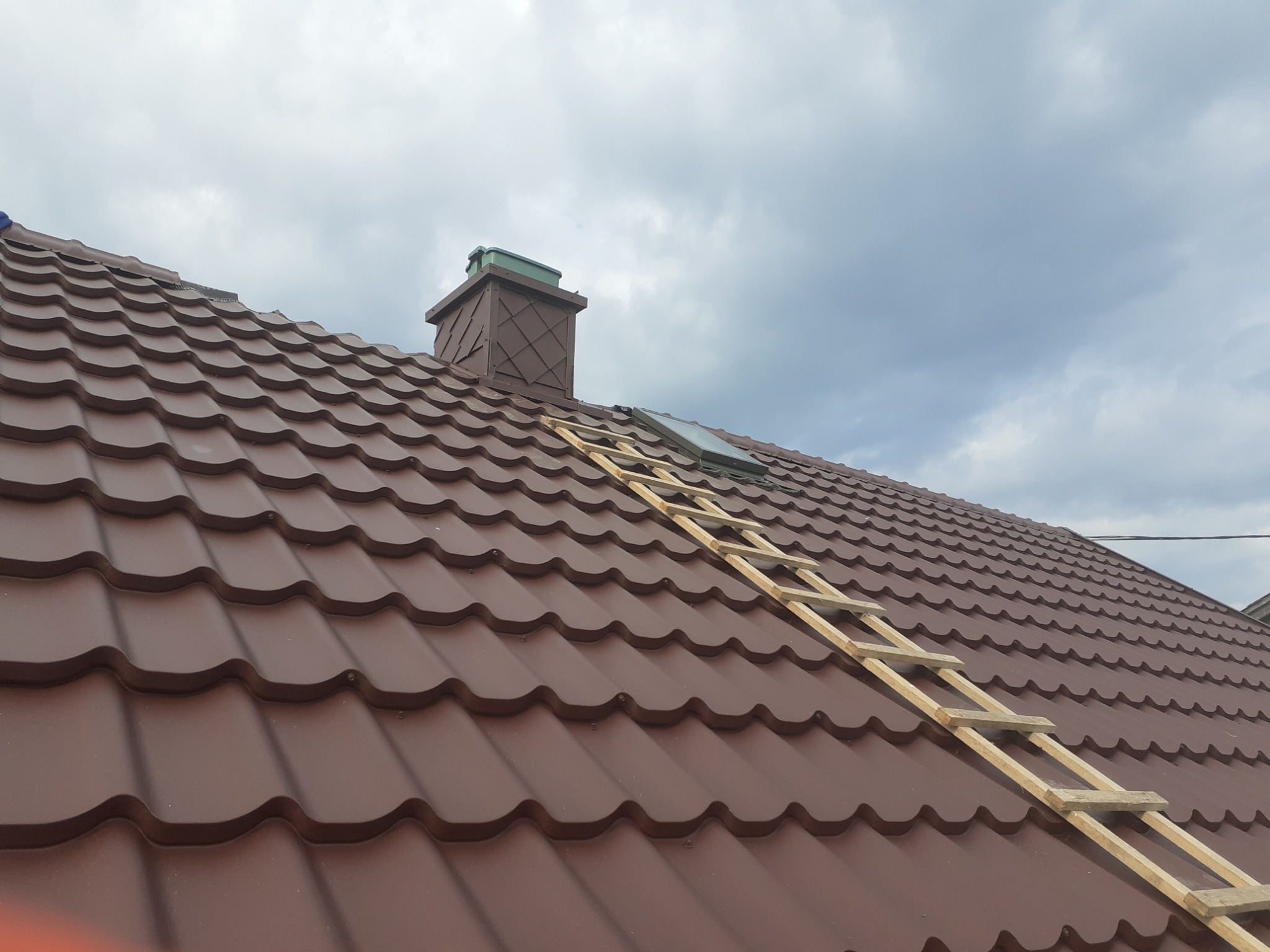 Brązowy dach z blachodachówki z drewnianą drabiną opartą o jego powierzchnię, widoczny komin i okno dachowe, pochmurne niebo w tle.