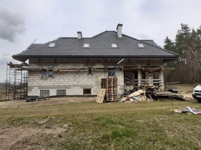 Dom w trakcie budowy z nowym, szarym dachem z dachówki ceramicznej, widoczne rusztowania i materiały budowlane na posesji.