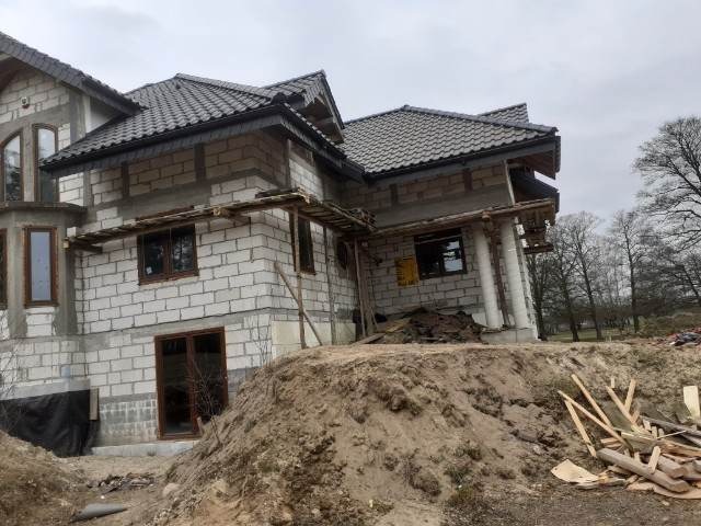 Dom w trakcie budowy z widoczną konstrukcją dachu i rusztowaniem, częściowo pokryty szarą dachówką, z hałdą ziemi i resztkami drewna na pierwszym planie.