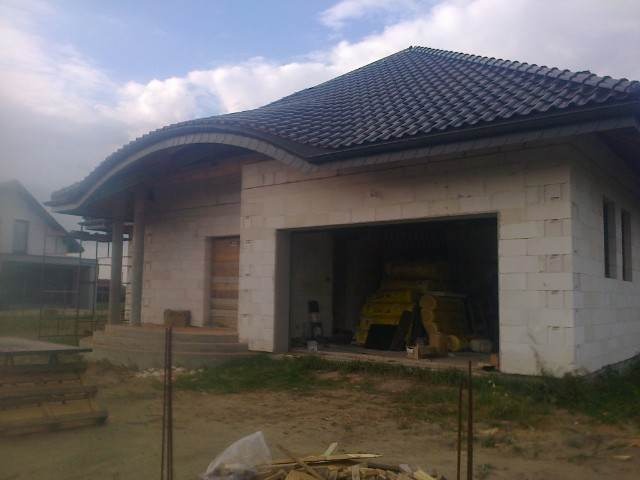 Budynek w trakcie budowy z widoczną konstrukcją dachu z czarnej dachówki, ściany z białych bloczków, widoczna więźba dachowa w garażu, niezagospodarowany teren wokół.