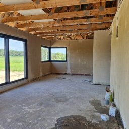 ZEN-TYNK - Surowe wnętrze domu w trakcie prac wykończeniowych: ściany po nałożeniu tynku, drewniana konstrukcja dachu, betonowa wylewka na podłodze z widocznymi śladami wilgoci, dwa okna z widokiem na zielone...