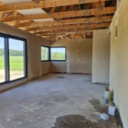 Surowe wnętrze domu w trakcie prac wykończeniowych: ściany po nałożeniu tynku, drewniana konstrukcja dachu, betonowa wylewka na podłodze z widocznymi śladami wilgoci, dwa okna z widokiem na zielone...