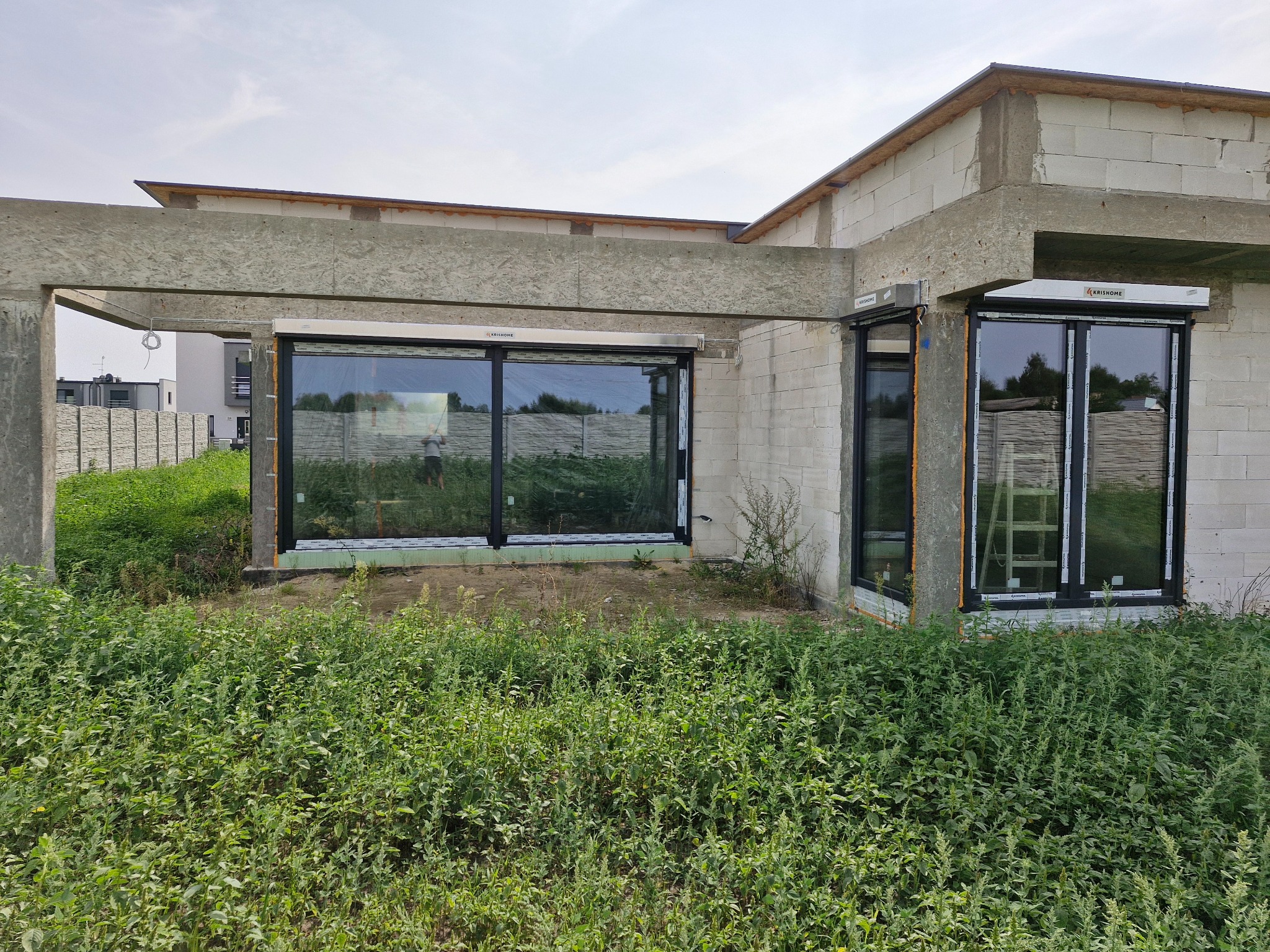 Surowy stan budynku z betonu komórkowego z zamontowanymi oknami z czarnymi ramami i widocznym ociepleniem wokół nich, otoczony wysoką trawą i chwastami.