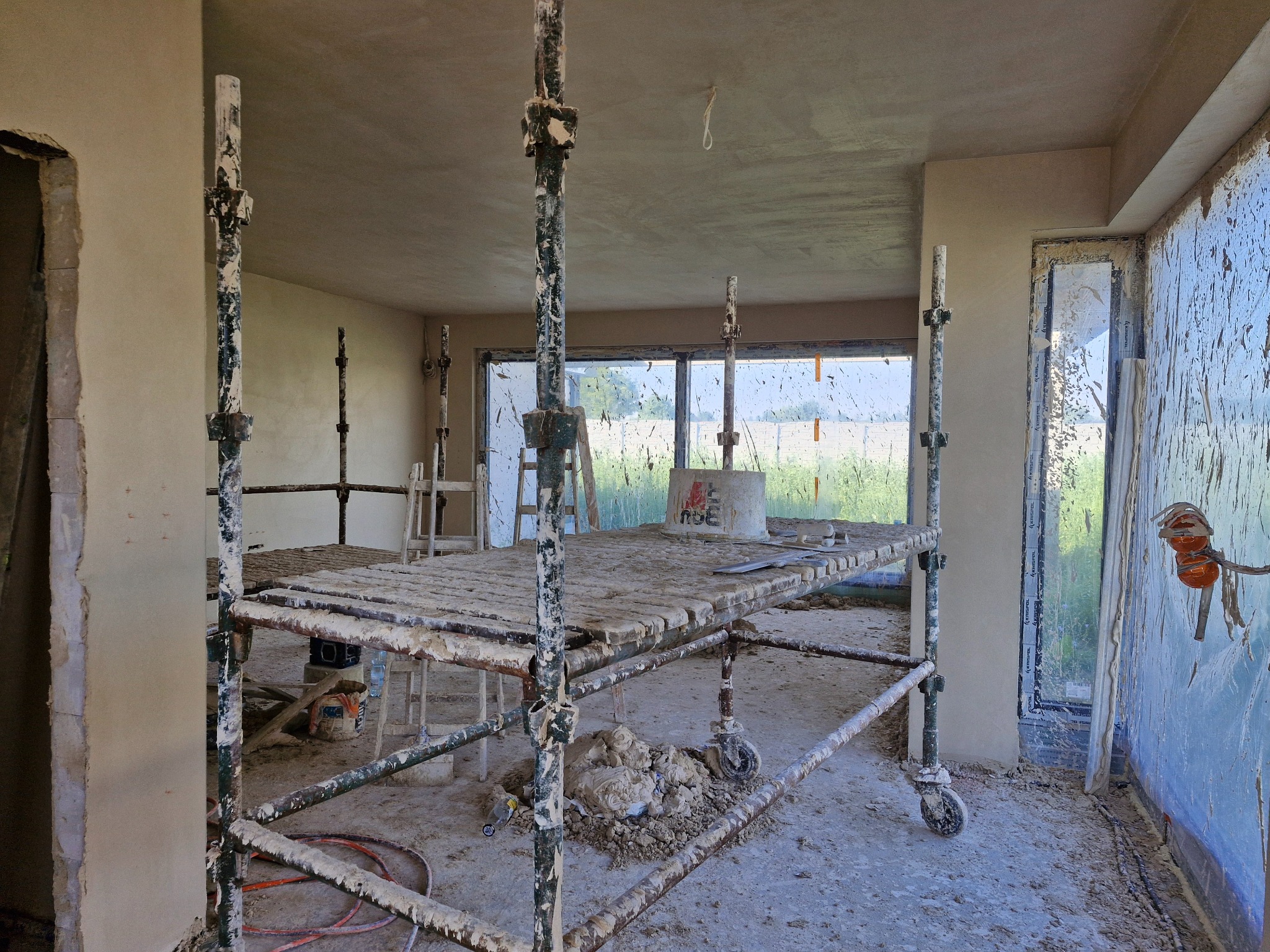 Wnętrze w trakcie tynkowania maszynowego: widoczne rusztowanie z resztkami tynku, świeżo otynkowane ściany, okna zabezpieczone folią, oraz wiadro z zaprawą na podłodze.