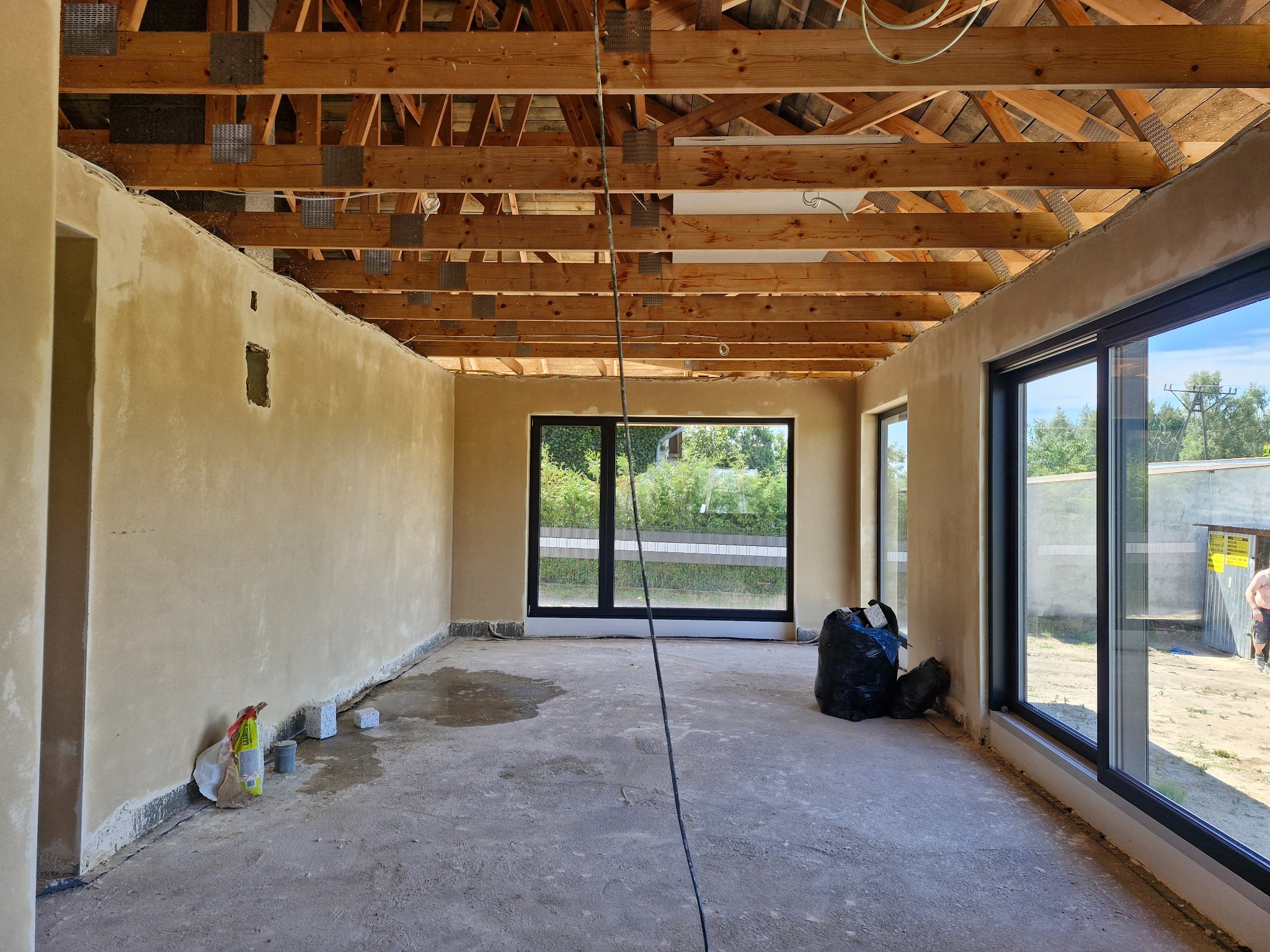 Surowe wnętrze w trakcie prac wykończeniowych: ściany pokryte wstępną warstwą tynku, widoczna konstrukcja drewniana dachu, duże okna z czarnymi ramami, na podłodze resztki materiałów budowlanych...