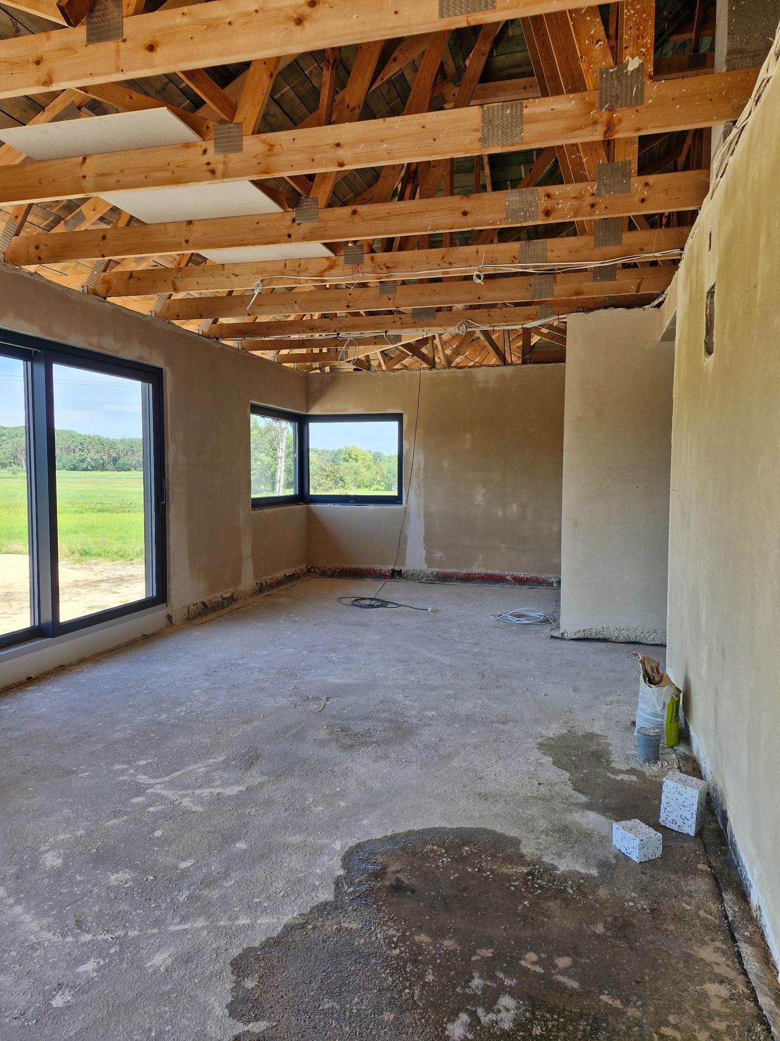 Surowe wnętrze domu w trakcie prac wykończeniowych: ściany po nałożeniu tynku, drewniana konstrukcja dachu, betonowa wylewka na podłodze z widocznymi śladami wilgoci, dwa okna z widokiem na zielone...