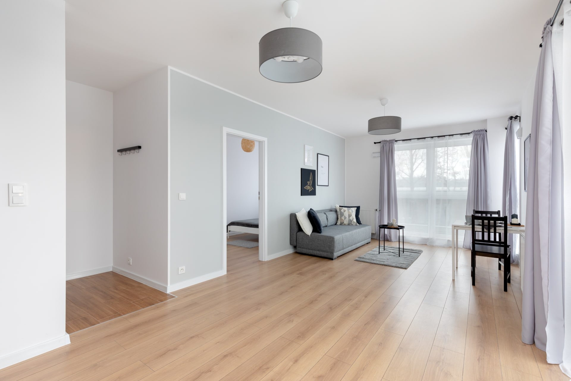 Jasne, przestronne wnętrze mieszkania z drewnianą podłogą, szarą sofą, stolikiem kawowym, zasłonami i widokiem na sypialnię w tle.
