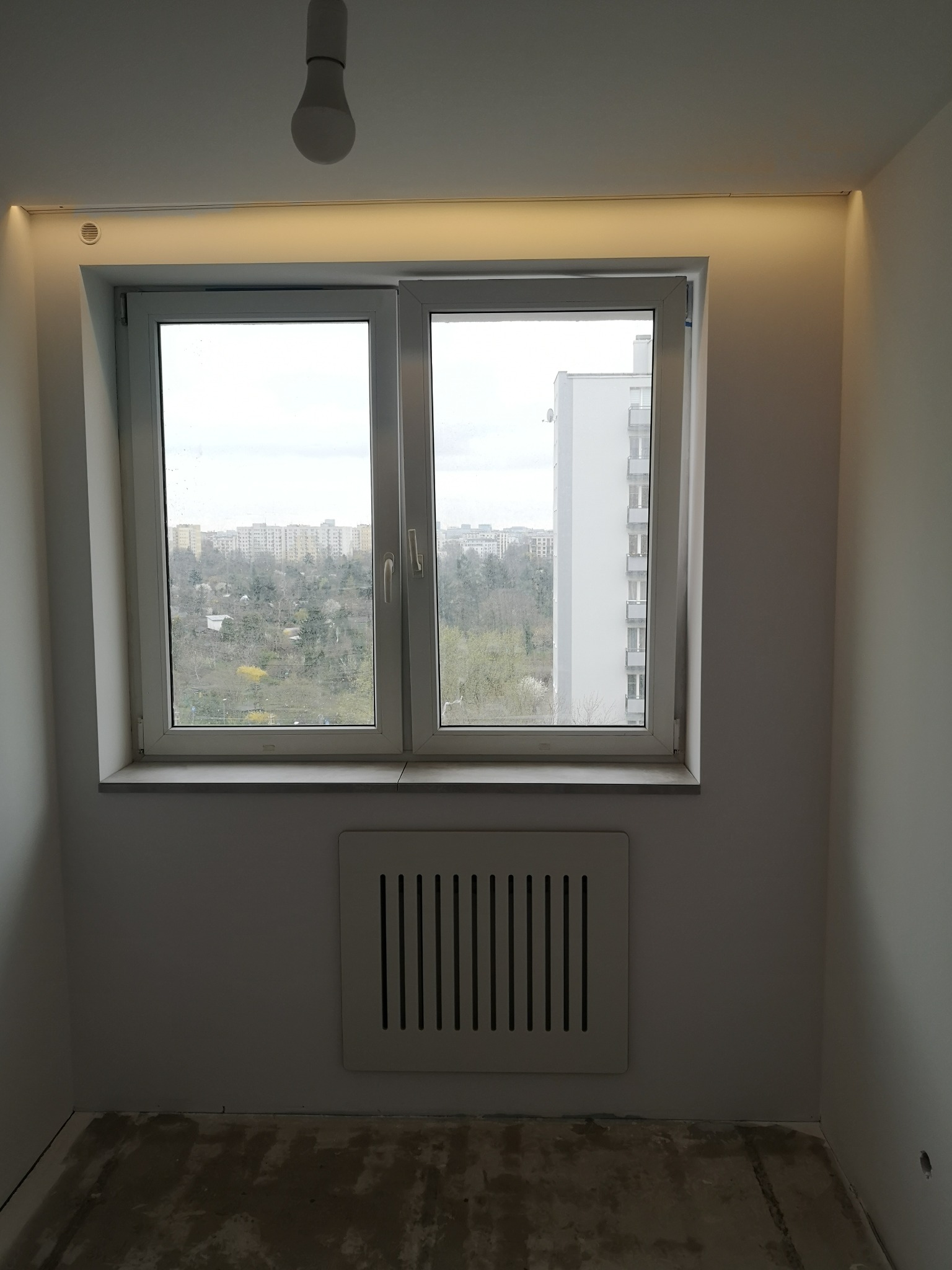 Pokój po remoncie z oknem, widok na miasto, podświetlony sufit LED, surowa podłoga i grzejnik z kratką.