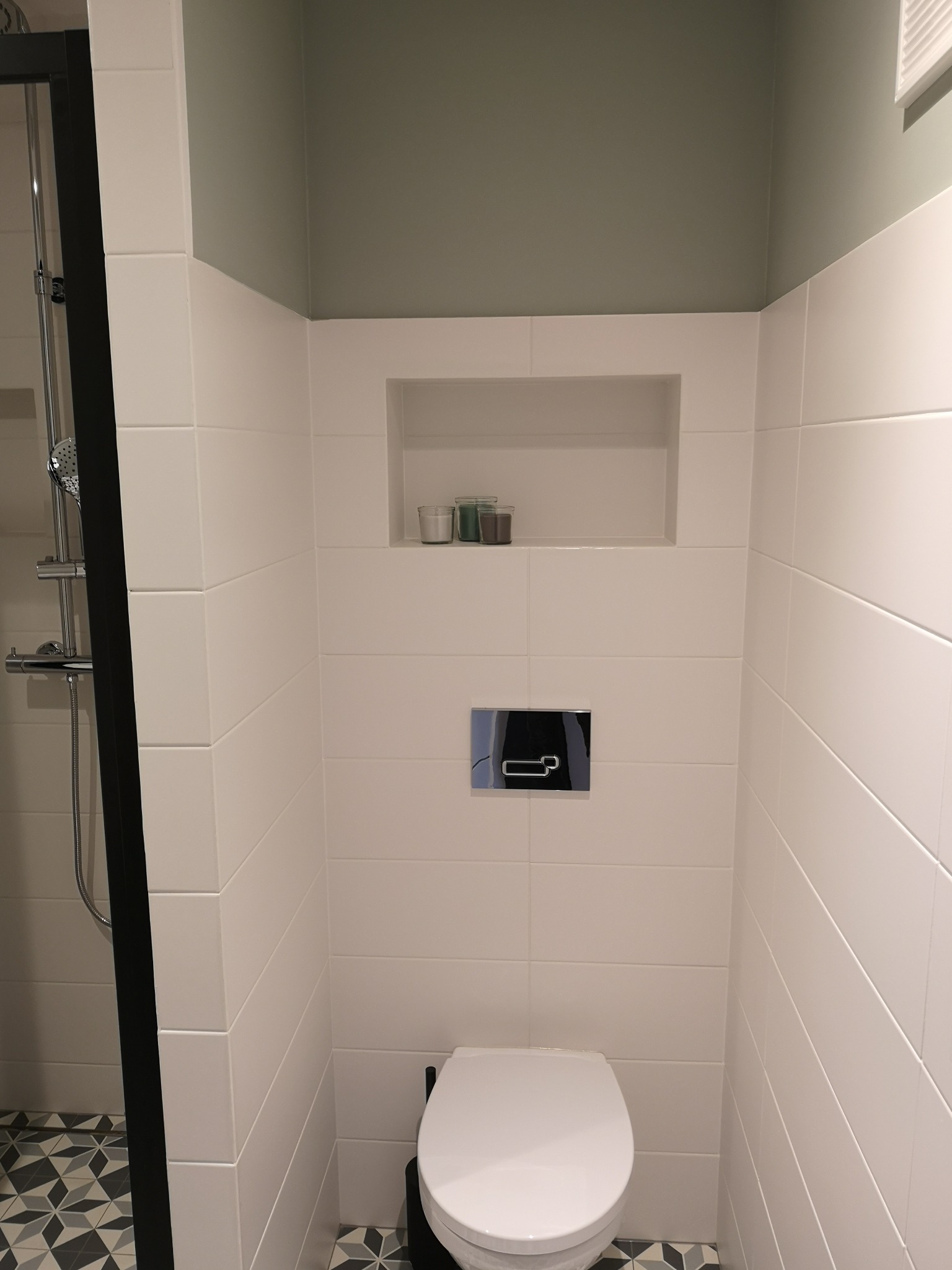 Wnętrze łazienki z białymi, poziomymi płytkami, wnęką z dekoracjami, toaletą i fragmentem kabiny prysznicowej z czarną ramą.