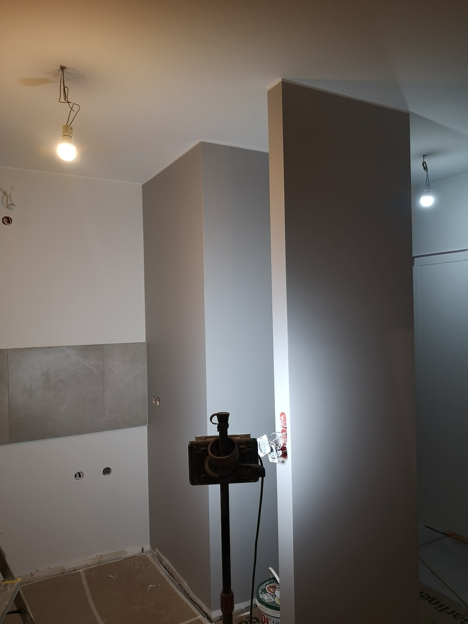 Świeżo pomalowane ściany w odcieniach szarości i bieli w trakcie remontu, widoczne przygotowane gniazdka elektryczne, fragment ułożonej glazury, lampa warsztatowa na statywie i drabina malarska.