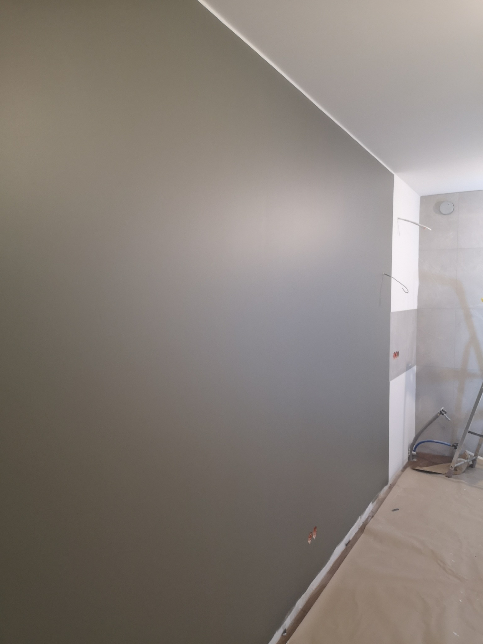 Świeżo pomalowana szara ściana w pomieszczeniu w trakcie remontu, widoczne wystające kable i przykryta podłoga.