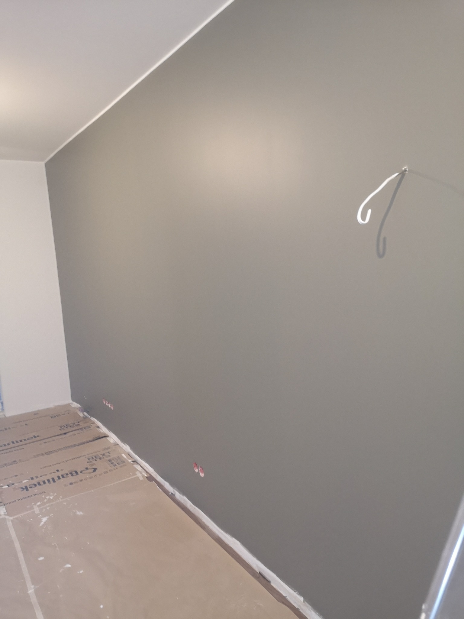 Świeżo pomalowana szara ściana z widocznymi puszkami elektrycznymi i zabezpieczoną podłogą kartonem, widoczne białe listwy przypodłogowe i sufitowe.