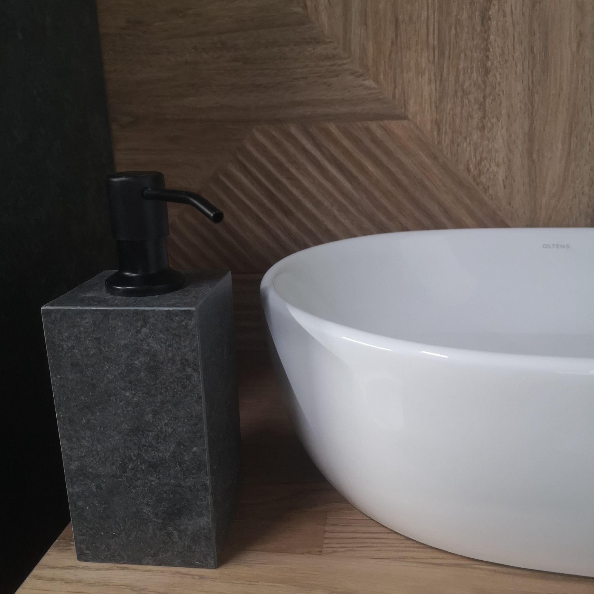 Biała umywalka nablatowa marki Oltens obok dozownika na mydło z szarego kamienia na drewnianym blacie, z dekoracyjną drewnianą ścianą w tle.