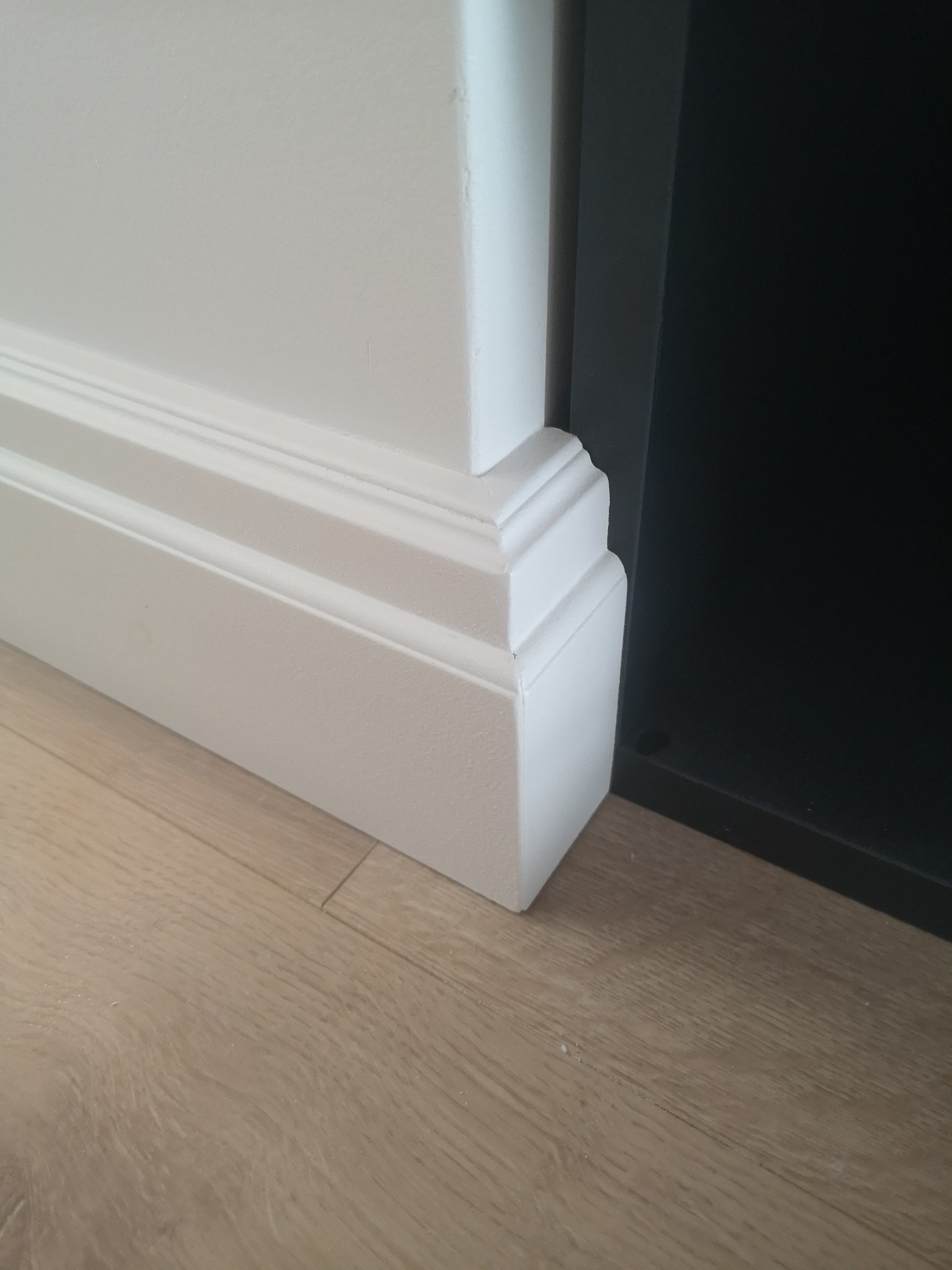 Biała listwa przypodłogowa z dekoracyjnym frezem, zamontowana przy ścianie pomalowanej na biało, na podłodze z paneli w odcieniach beżu i brązu, obok ciemnego mebla.