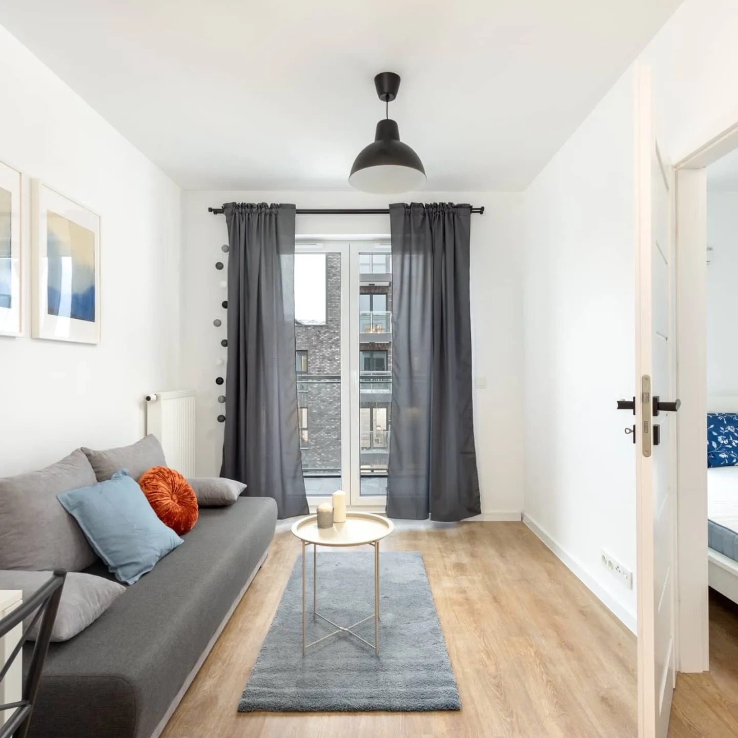 Jasne wnętrze pokoju dziennego z szarą sofą, błękitną i pomarańczową poduszką, stolikiem kawowym na dywanie oraz drewnianą podłogą, widok na budynek za oknem.