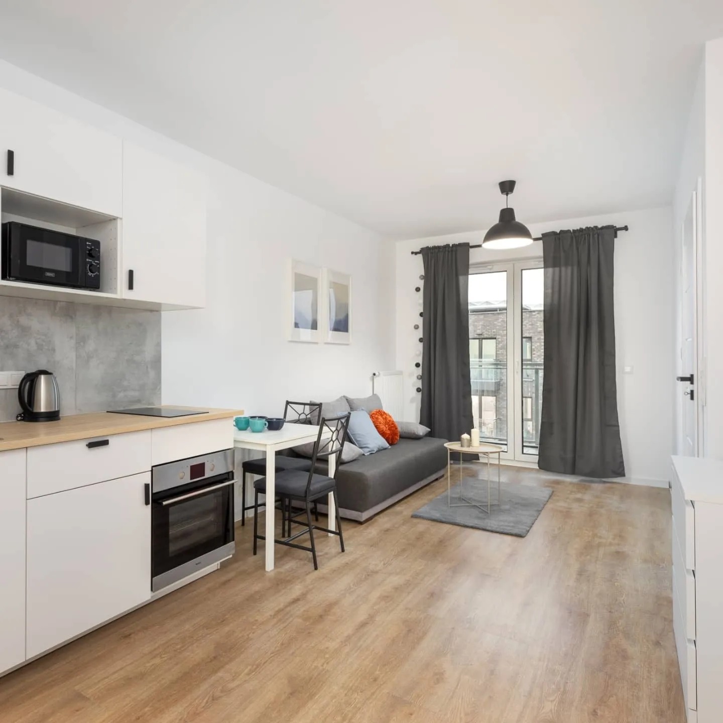 Jasne wnętrze mieszkania z aneksem kuchennym, białymi meblami, drewnianą podłogą i szarą sofą z kolorowymi poduszkami, widziane z niskiego kąta.