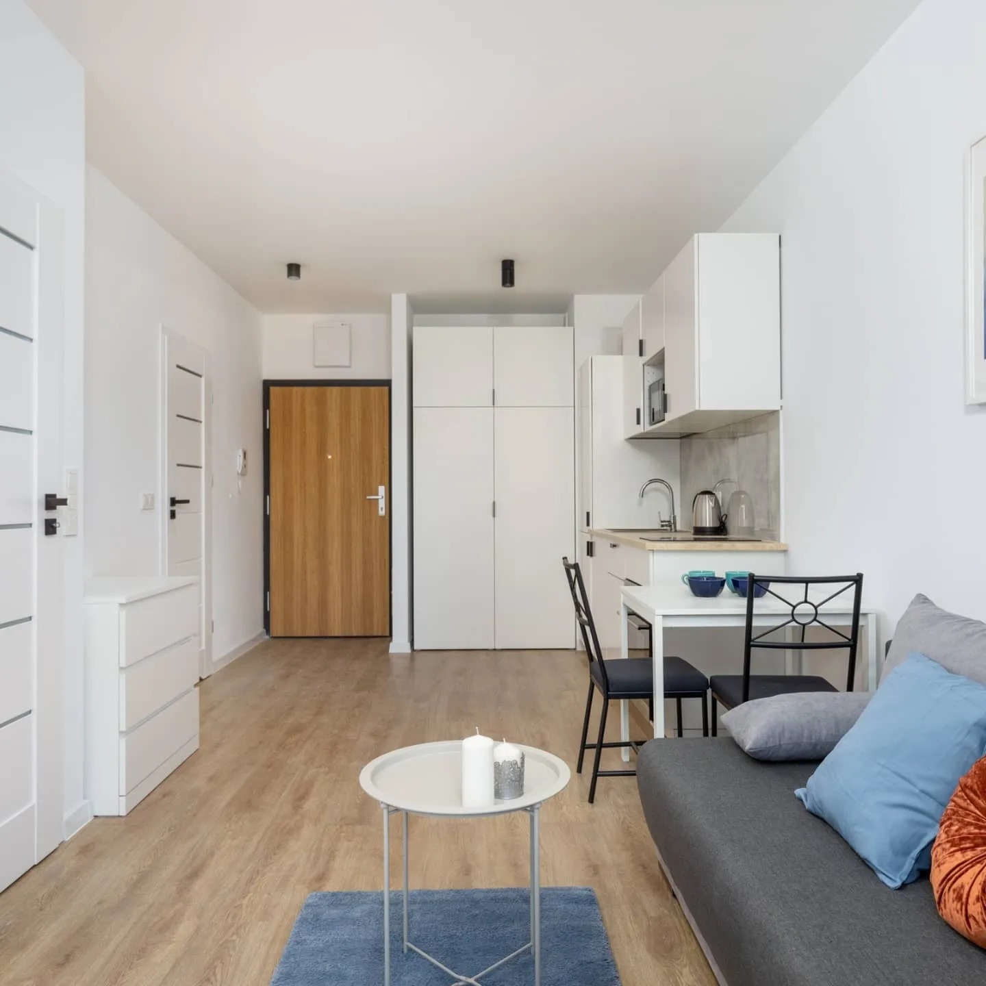 Jasne, minimalistyczne wnętrze małego mieszkania z aneksem kuchennym, drewnianą podłogą i akcentami w odcieniach błękitu i szarości, widok na drzwi wejściowe.