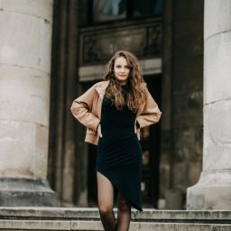 Fotoleśna - Martyna Majda - Młoda kobieta z kręconymi włosami w czarnej sukience i beżowej kurtce pozuje na kamiennych schodach przed budynkiem z kolumnami.