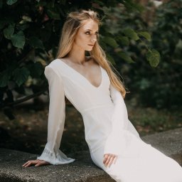 Fotoleśna - Martyna Majda - Elegancka blondynka w białej sukni z szerokimi rękawami, pozuje siedząc na kamiennym murku na tle zielonych liści.
