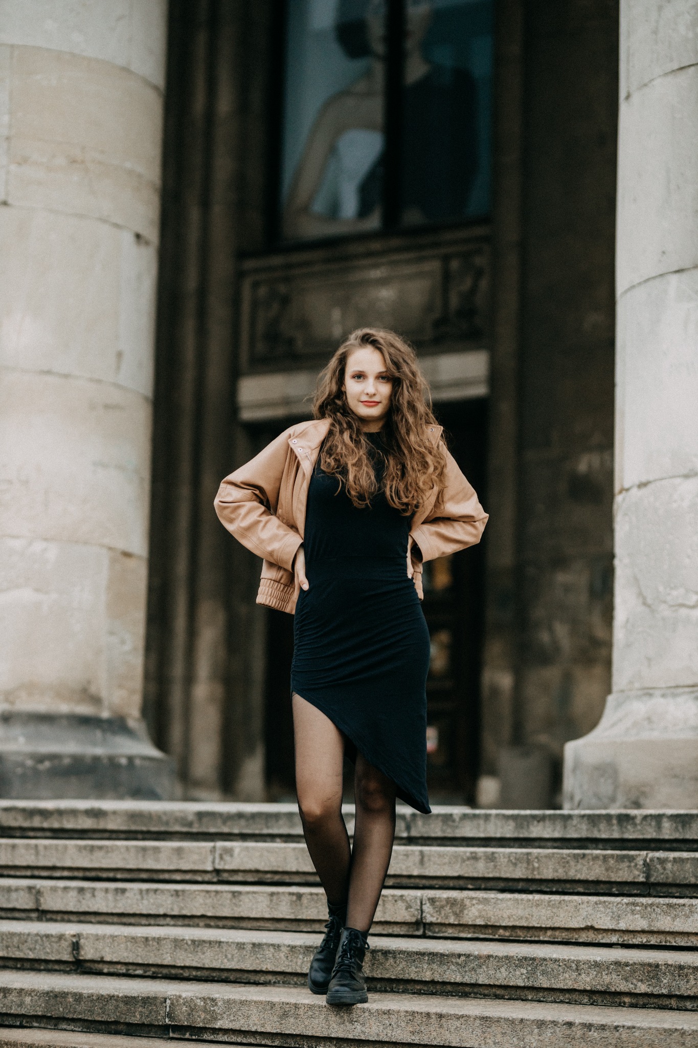 Młoda kobieta z kręconymi włosami w czarnej sukience i beżowej kurtce pozuje na kamiennych schodach przed budynkiem z kolumnami.
