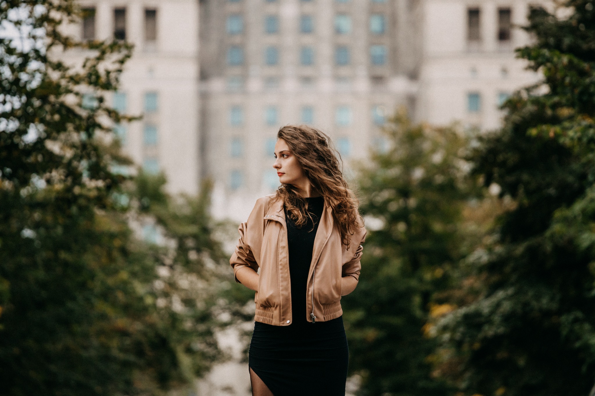 Kobieta w czarnej sukience i beżowej kurtce pozuje na tle budynku z wieloma oknami, otoczona zielenią.