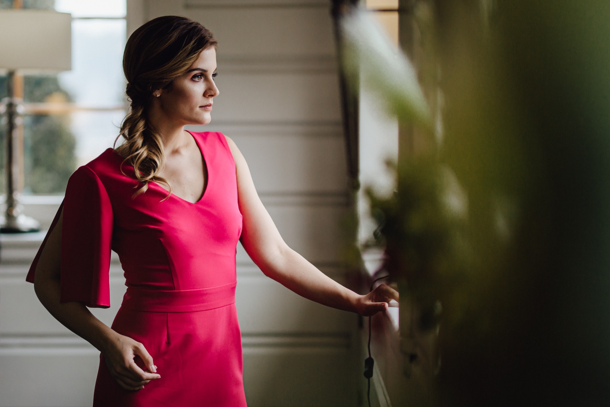 Kobieta w różowej sukience z rozciętymi rękawami, spogląda przez okno, z rozpuszczonymi włosami spiętymi w warkocz.