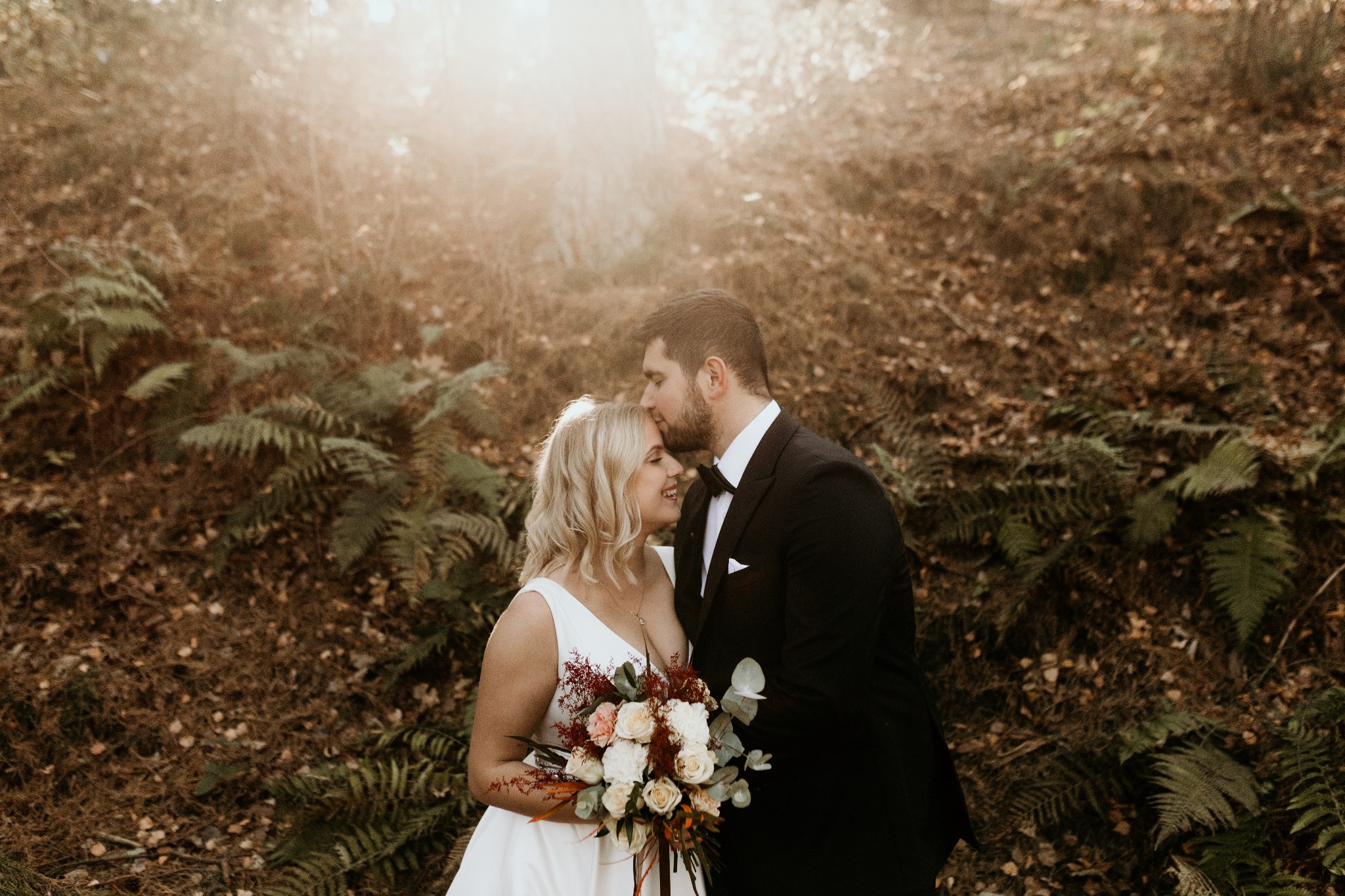 Para młoda w lesie: panna młoda w białej sukni trzyma bukiet, pan młody w czarnym smokingu całuje ją w czoło na tle paproci i opadłych liści, ciepłe światło słoneczne