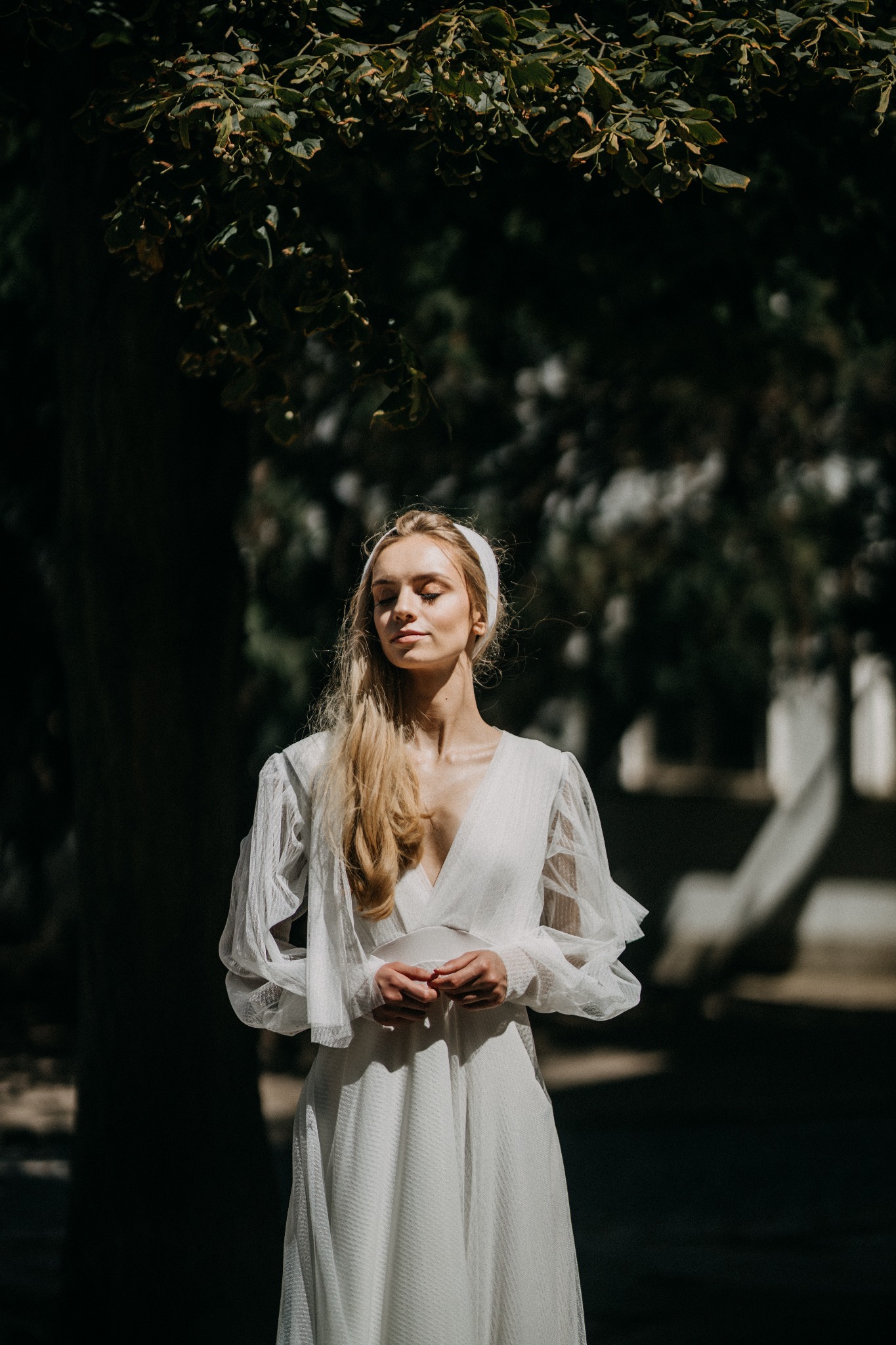 Kobieta w białej sukni z bufiastymi rękawami, z zamkniętymi oczami, stojąca w cieniu drzew w Warszawie, trzymająca dłonie blisko siebie.