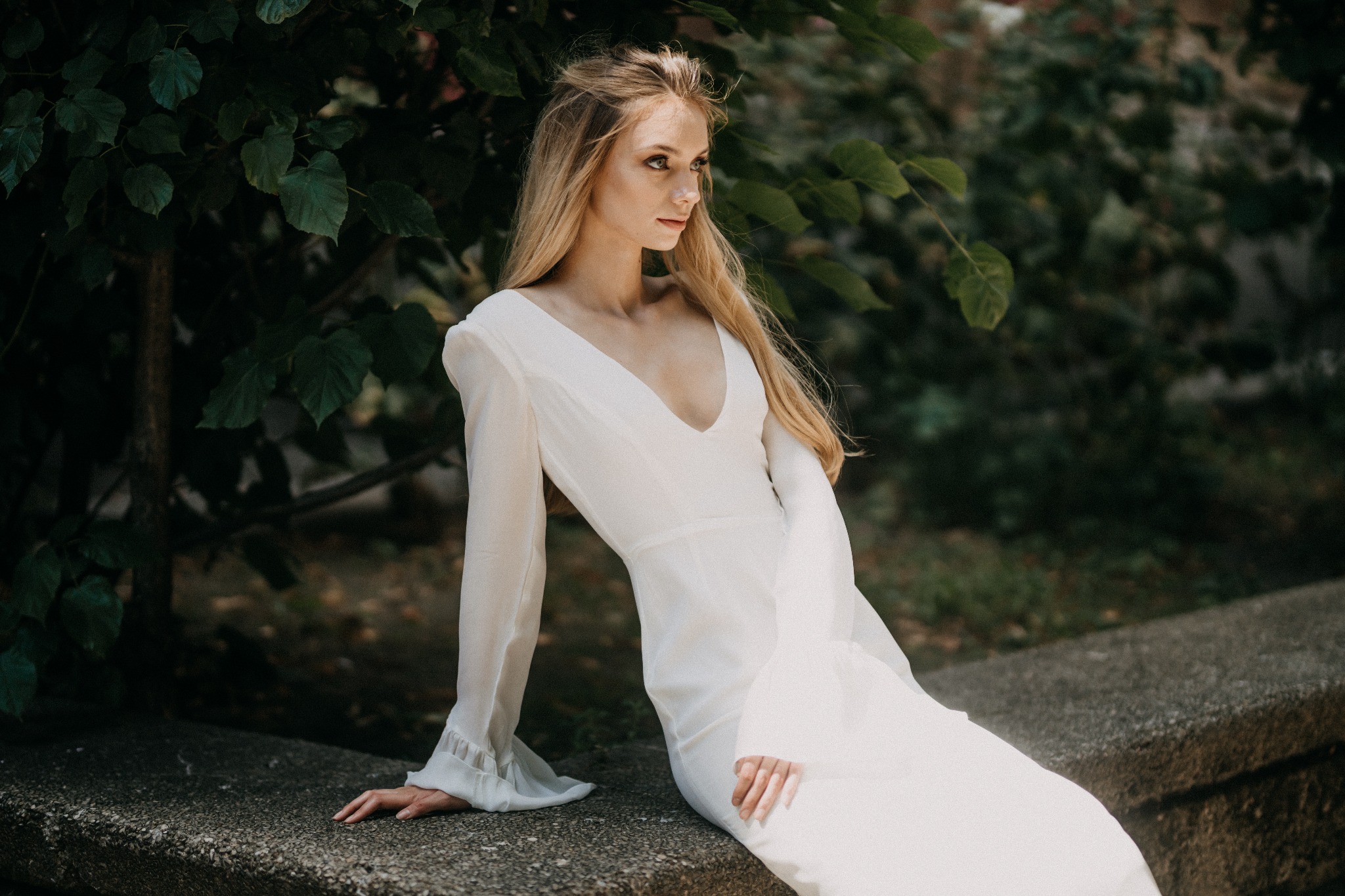 Elegancka blondynka w białej sukni z szerokimi rękawami, pozuje siedząc na kamiennym murku na tle zielonych liści.