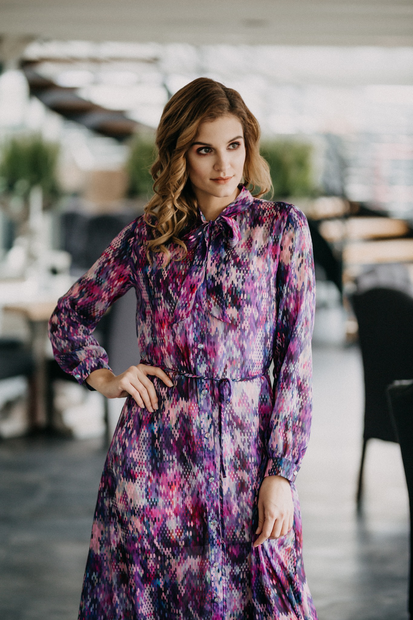 Kobieta w długiej, wzorzystej sukience z wiązaniem pod szyją, pozująca z ręką na biodrze w jasnym wnętrzu.