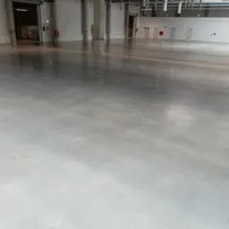 Błyszcząca, gładka posadzka w hali magazynowej lub produkcyjnej po czyszczeniu, widoczne refleksy świetlne, w tle otwarte wrota i drzwi.