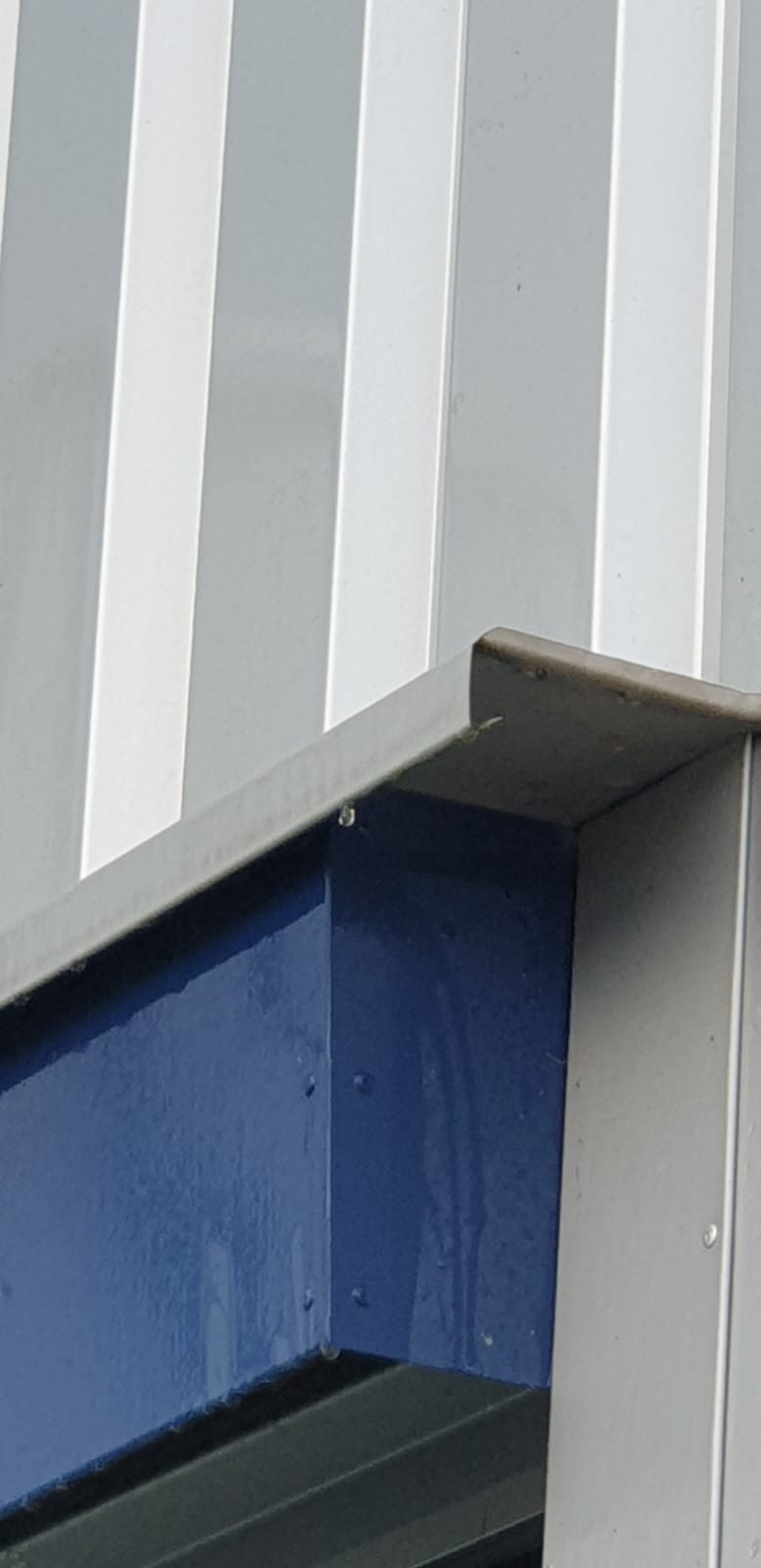 Detal zewnętrzny budynku przemysłowego z widocznymi śladami wilgoci po niedawnym myciu, krople wody na pomalowanej na niebiesko konstrukcji stalowej.