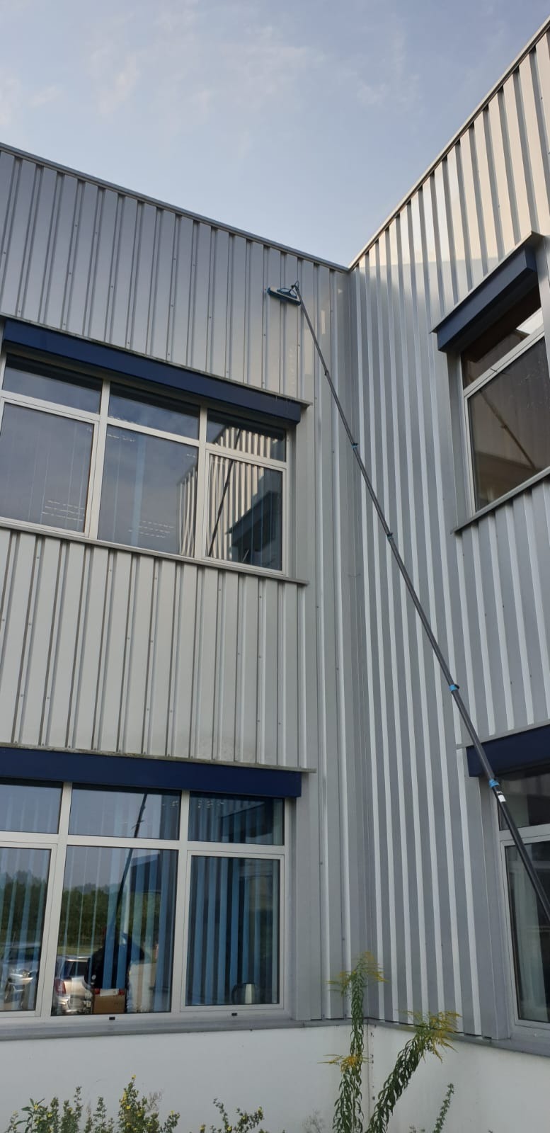 Mycie okien na wysokościach przy użyciu teleskopowego kija w Pawłowiczkach. Budynek biurowy z szarymi panelami elewacyjnymi i niebieskimi ramami okiennymi.