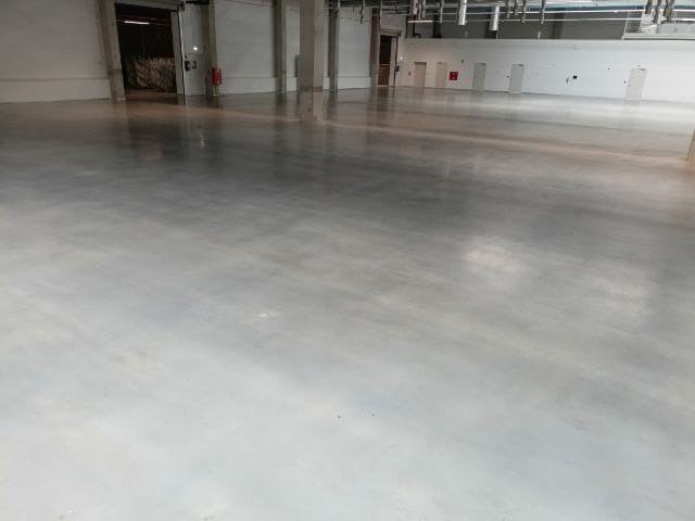 Błyszcząca, gładka posadzka w hali magazynowej lub produkcyjnej po czyszczeniu, widoczne refleksy świetlne, w tle otwarte wrota i drzwi.