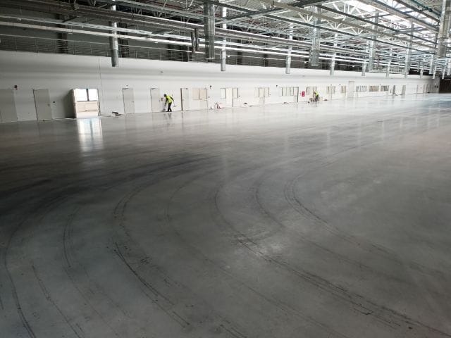 Duża, pusta hala przemysłowa z betonową podłogą i pracownikami w odblaskowych kamizelkach przy ścianie z rzędem drzwi.