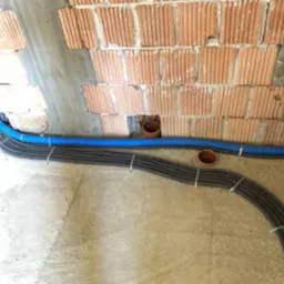 Wiązka ciemnych, karbowanych przewodów w peszlu, mocowanych metalowymi uchwytami do surowej, betonowej podłogi i ceglanej ściany w trakcie prac instalacyjnych.