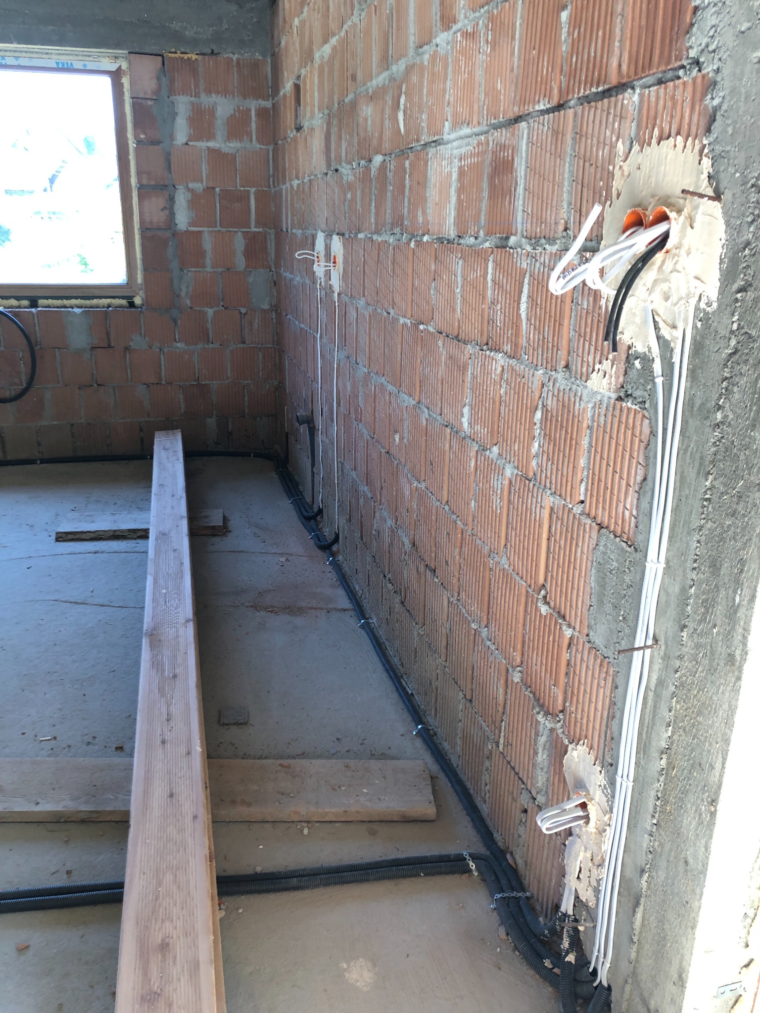 Surowy stan budynku w trakcie prac elektrycznych: widoczne kable w peszlach i przewodach mocowane do ściany z cegły, otwory w ścianie wypełnione pianką montażową, oraz deska na betonowej podłodze.