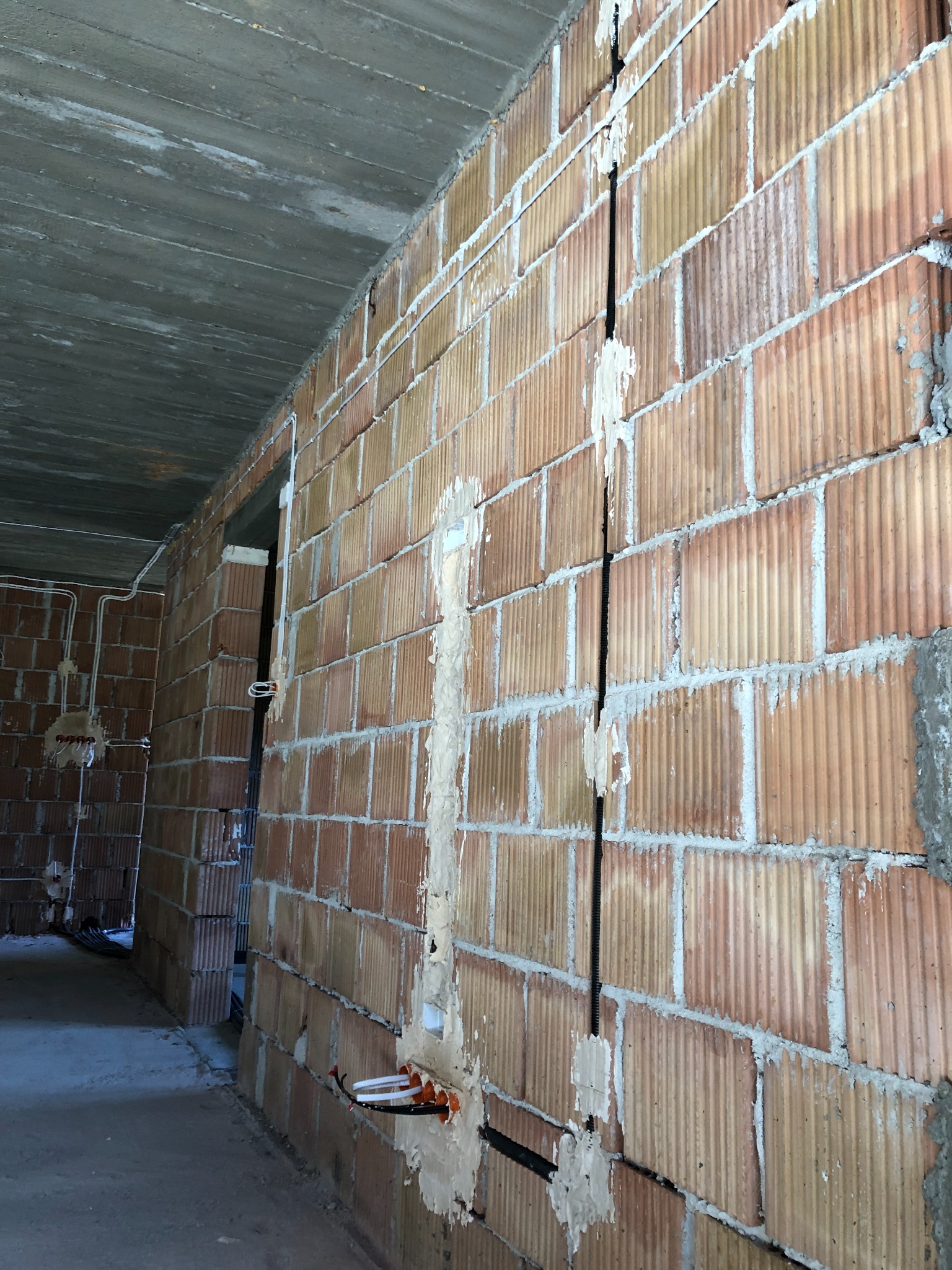 Wewnętrzna ściana z czerwonej cegły z wyżłobionymi kanałami, w których poprowadzone są czarne rury instalacyjne; widoczne puszki instalacyjne z przewodami.