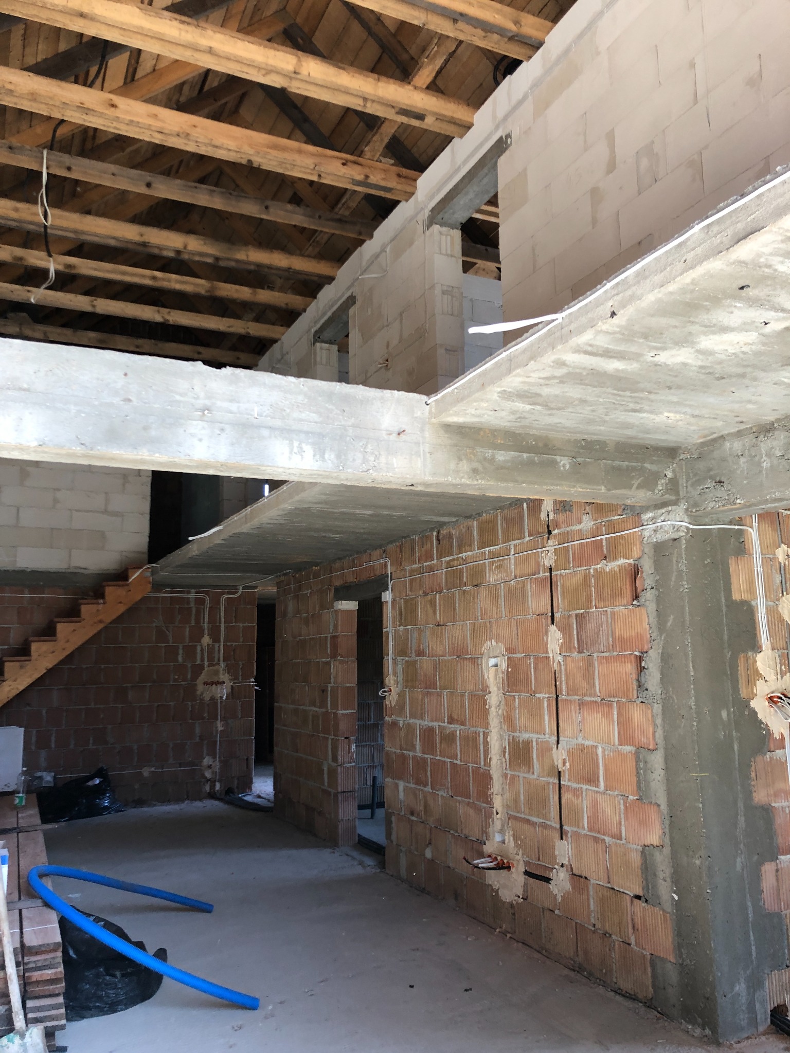 Wnętrze surowego domu w trakcie budowy, widoczne ceglane ściany z bruzdami pod instalacje, drewniana konstrukcja dachu oraz fragment schodów.