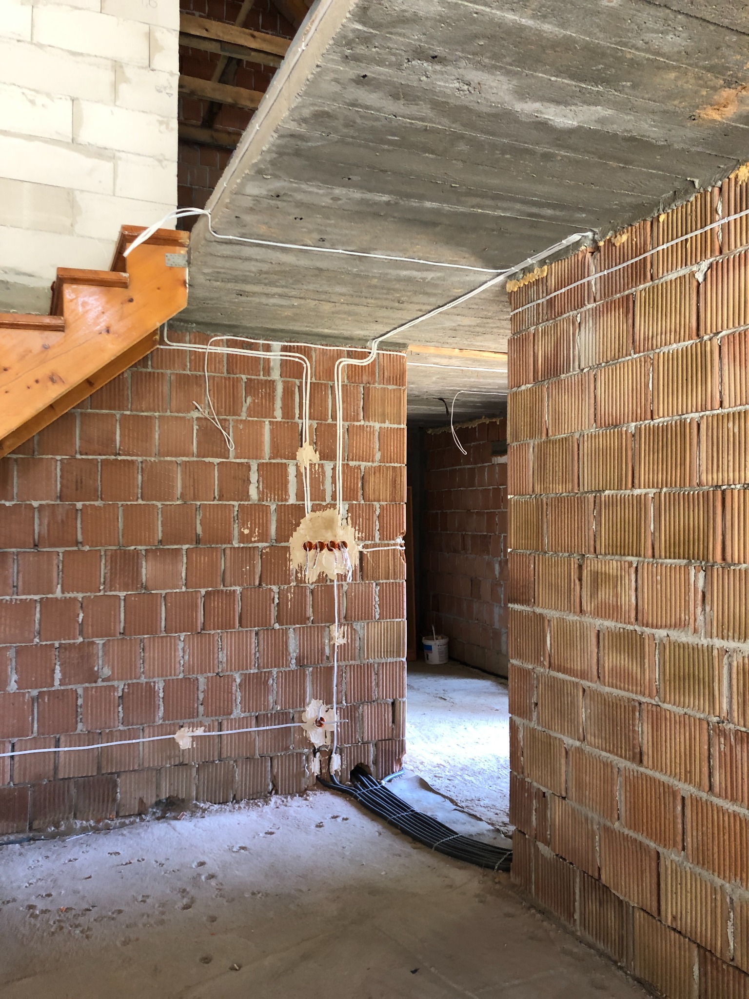Wnętrze w trakcie budowy z widocznymi instalacjami elektrycznymi: kable prowadzone po ścianach z cegły ceramicznej, puszki instalacyjne częściowo zamontowane, surowy betonowy strop, drewniane...