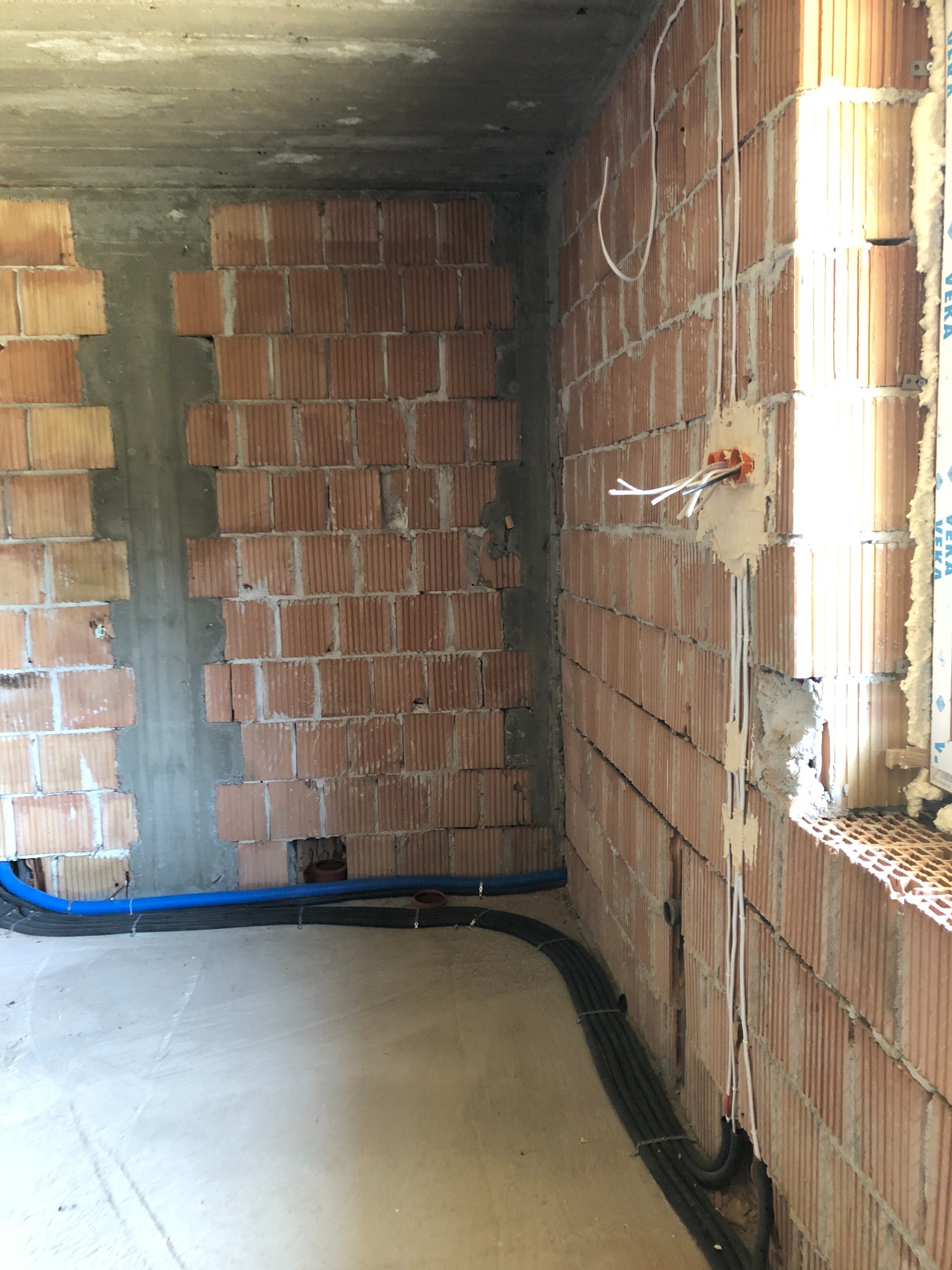 Surowy mur z cegły z widocznymi przewodami elektrycznymi, puszką instalacyjną oraz niebieską rurą karbowaną przy podłodze w trakcie prac instalacyjnych.