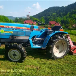 Niebieski traktor Kumiai MT1601D na trawiastym terenie z widokiem na zalesione wzgórza i wieżę w oddali.