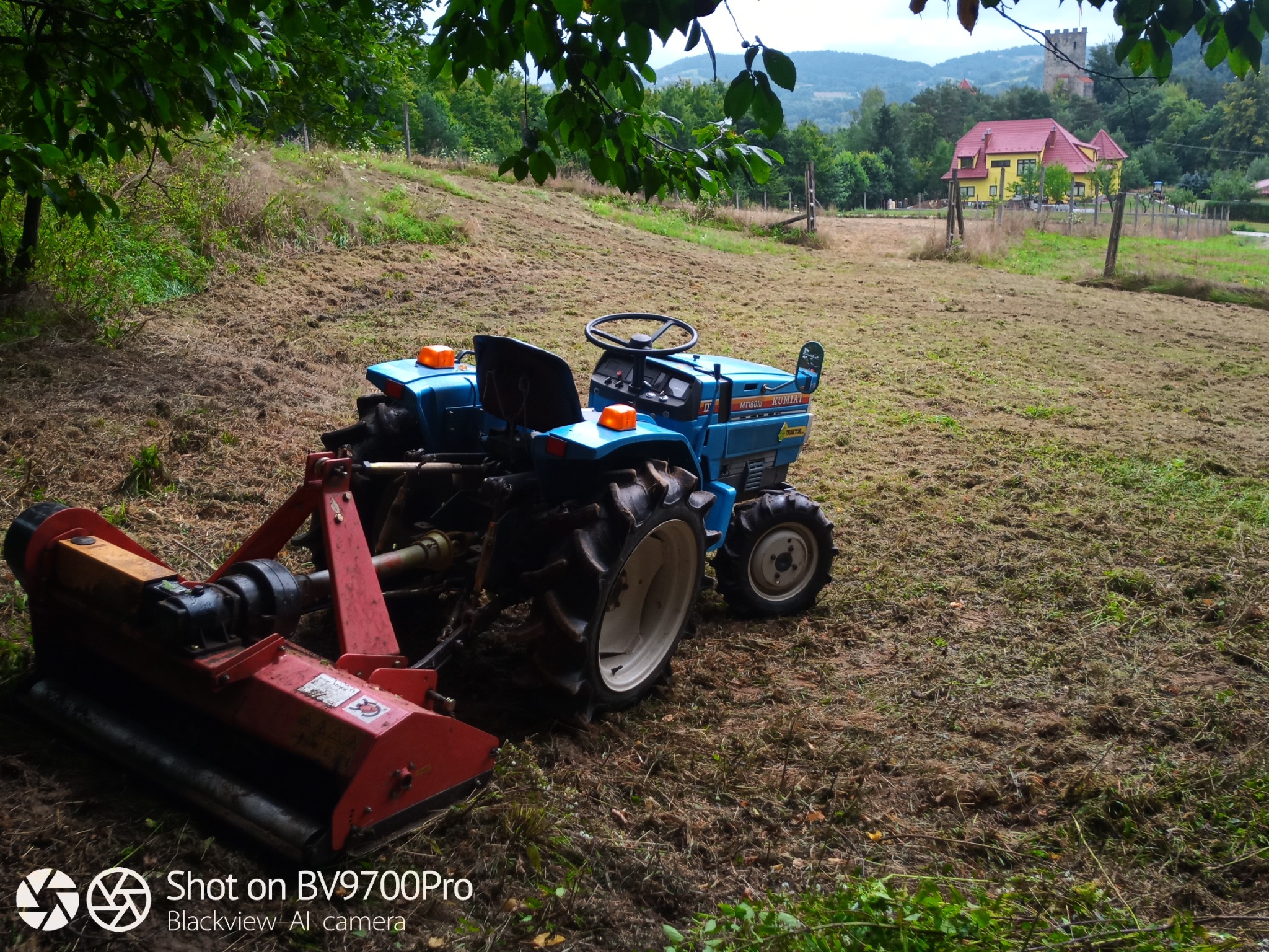 Niebieski traktor z czerwonym kosiarką rotacyjną na polu świeżo skoszonej trawy. W tle żółty dom z czerwonym dachem i wieża zamkowa na wzgórzu.