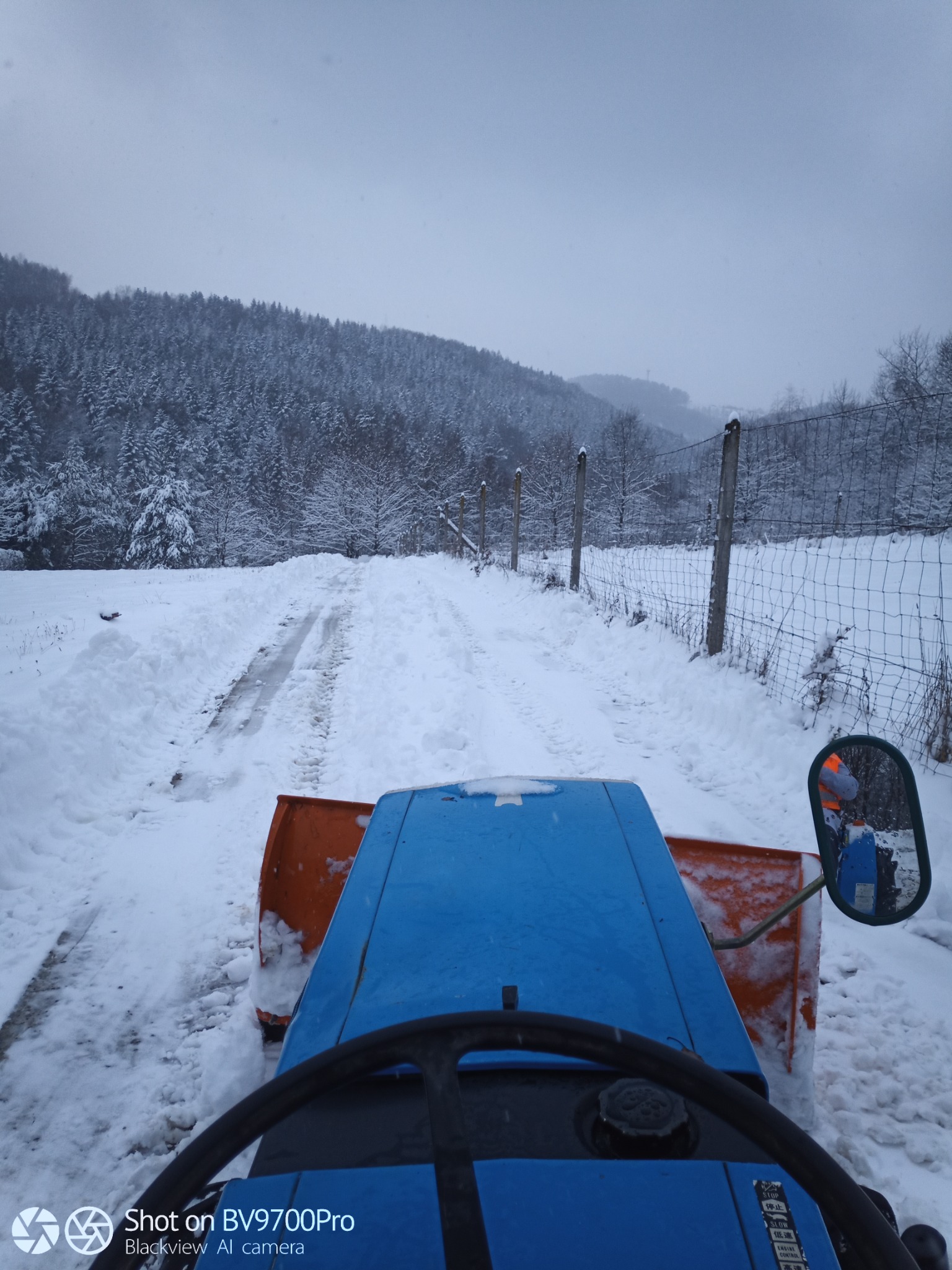 Widok z perspektywy kierowcy niebieskiego traktora z pomarańczowym pługiem odśnieżającym, jadącego po zasypanej śniegiem drodze w kierunku zalesionego wzgórza w pochmurny dzień.