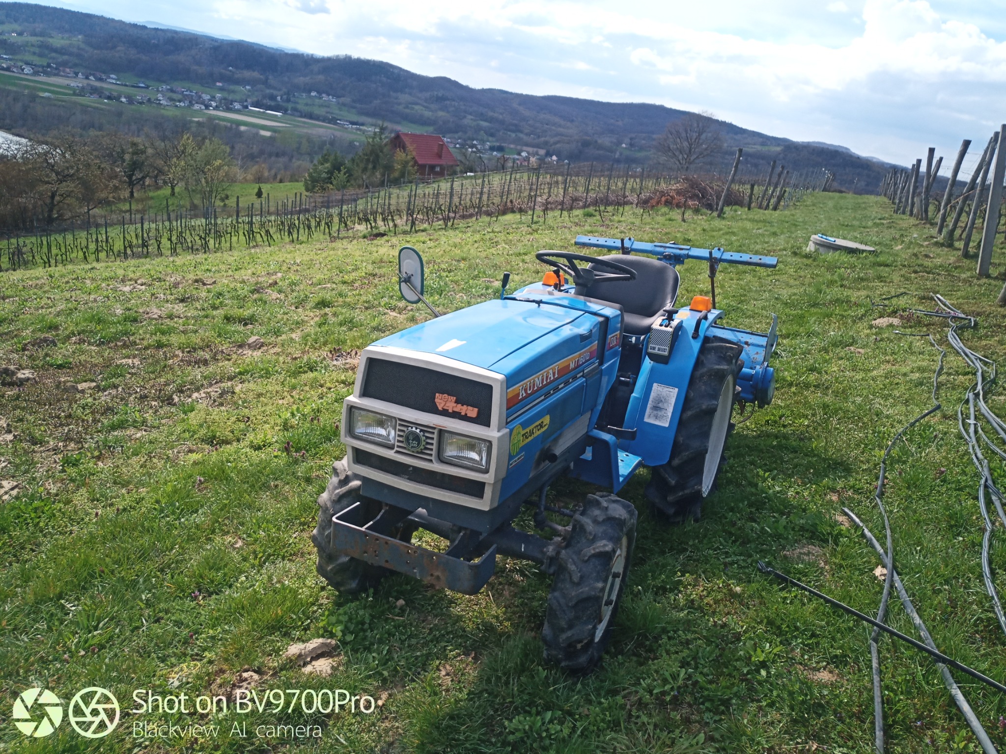 Niebieski traktor Kubota MT160D na nierównym terenie porośniętym trawą, w tle winnica, dom z czerwoną dachówką i wzgórza pod błękitnym niebem.