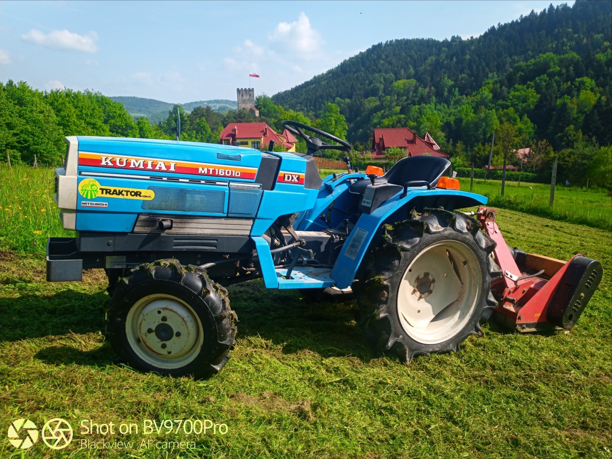 Niebieski traktor Kumiai MT1601D na trawiastym terenie z widokiem na zalesione wzgórza i wieżę w oddali.