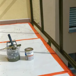 Malowanie balustrady balkonowej farbą Sigma, widoczny pędzel, wiadro z farbą i zabezpieczona folią podłoga. Świeżo pomalowane elementy na pomarańczowo.