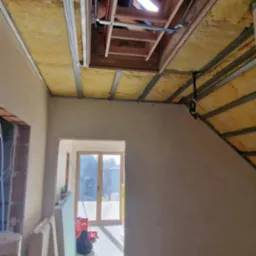 Ocieplone wełną poddasze z ramą drewnianą wokół okna dachowego i metalowym stelażem sufitu podwieszanego. Widok na wnętrze domu w trakcie prac wykończeniowych.